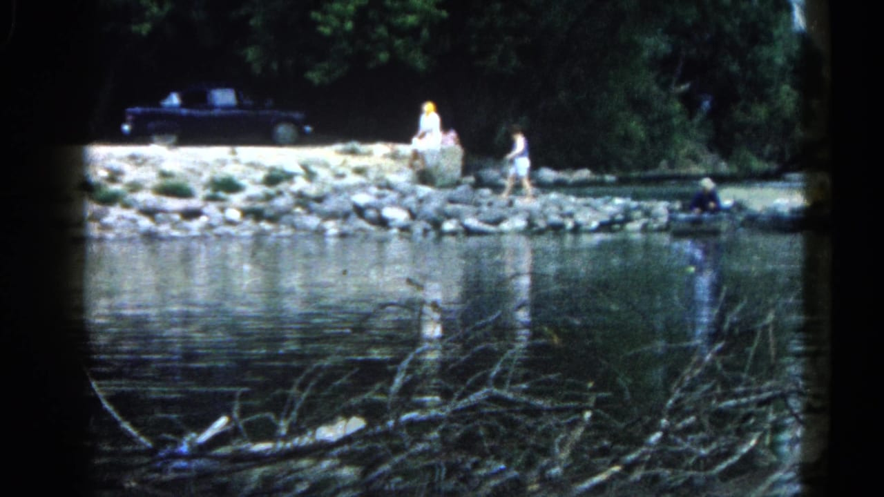 8mm home movie frame, c.1962 Illinois — two adults stroll a rocky riverbank in the middle distance; a large dark vintage car is parked under trees; branch debris floats in the foreground water.