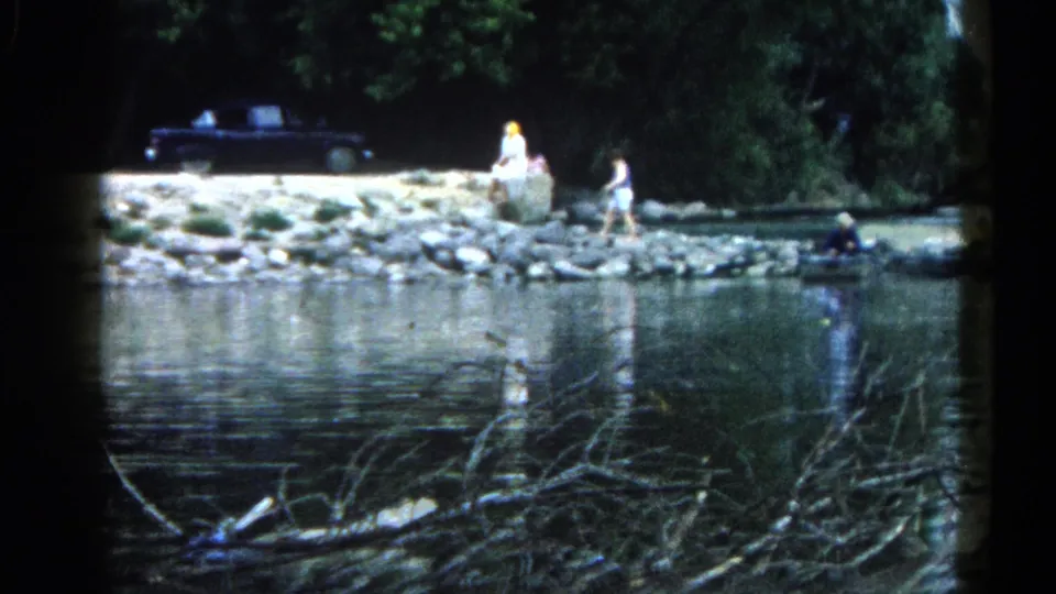8mm home movie frame, c.1962 Illinois — two adults stroll a rocky riverbank in the middle distance; a large dark vintage car is parked under trees; branch debris floats in the foreground water.