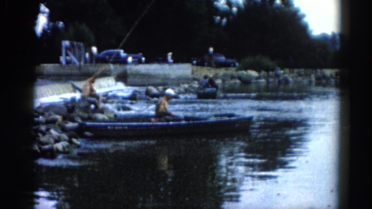 Two men fishing from a small boat and rocky riverbank, captured in early 1960s 8mm home movie footage.
