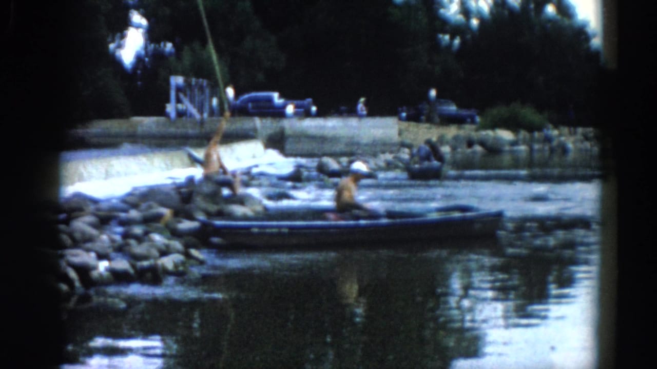 Man casting a fishing line from a small boat on a river; rocky shoreline and parked cars visible on the bank behind.