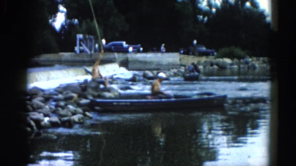 A man casting a fishing line from a small boat on an Illinois river, circa 1962; rocky breakwater and early-1960s automobiles visible on the upper bank.
