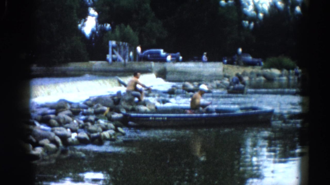 Two adult men fishing from a small boat and rocky shore on an Illinois river, circa 1962; early-1960s automobiles parked along the upper bank.