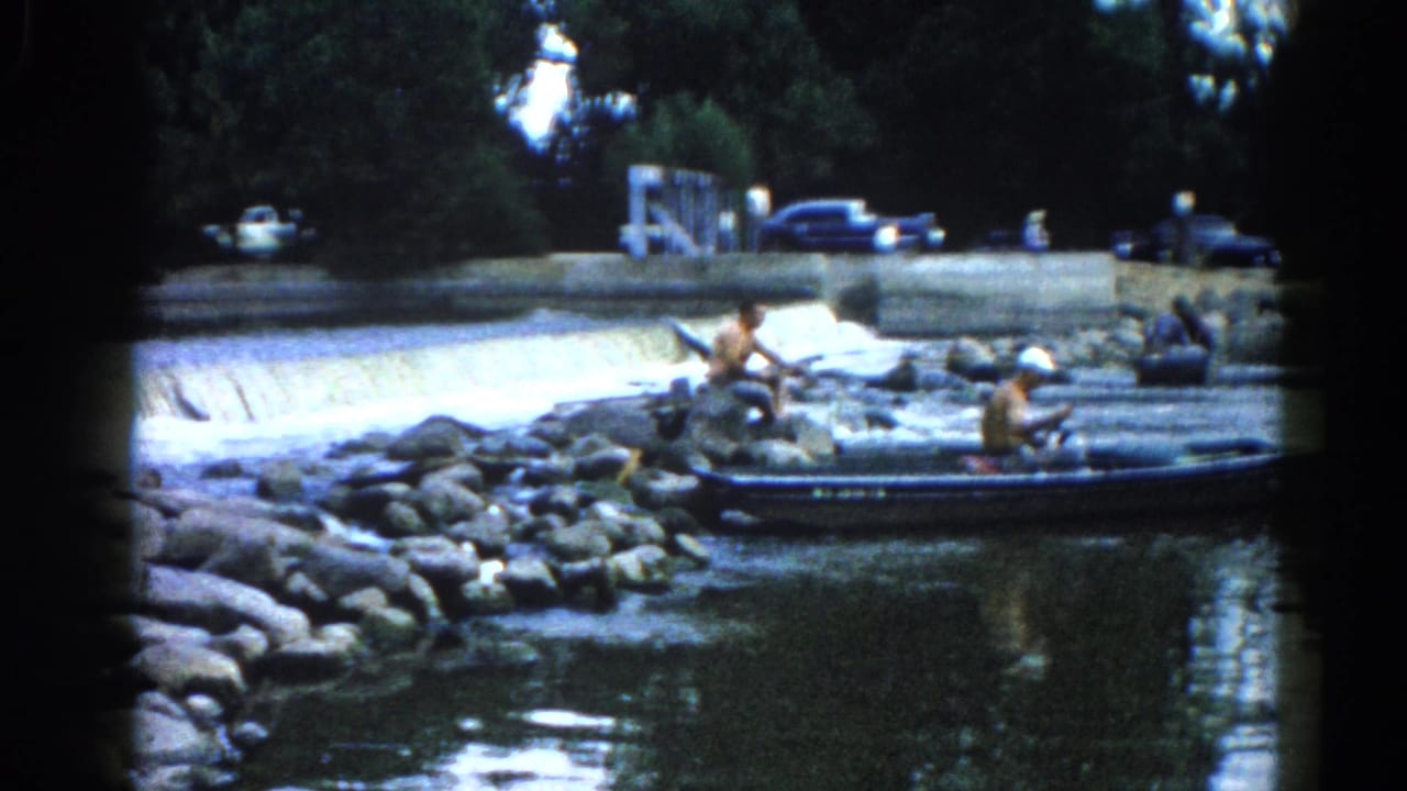 Two adult men fishing from a small boat and adjacent rocky shore on an Illinois river, circa 1962; dark-colored early-1960s automobiles visible on the upper bank.