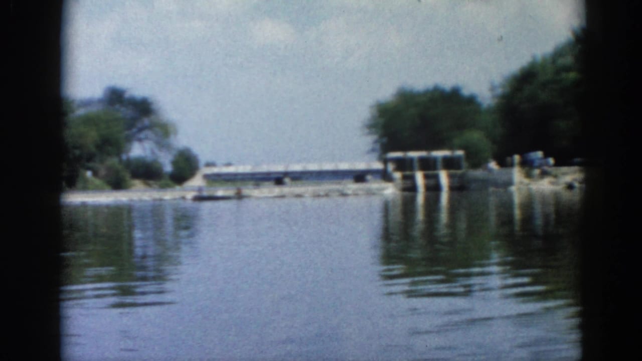 Wide view of a calm river with a concrete dam or lock structure on the far bank and tree-lined shores under a pale overcast sky.