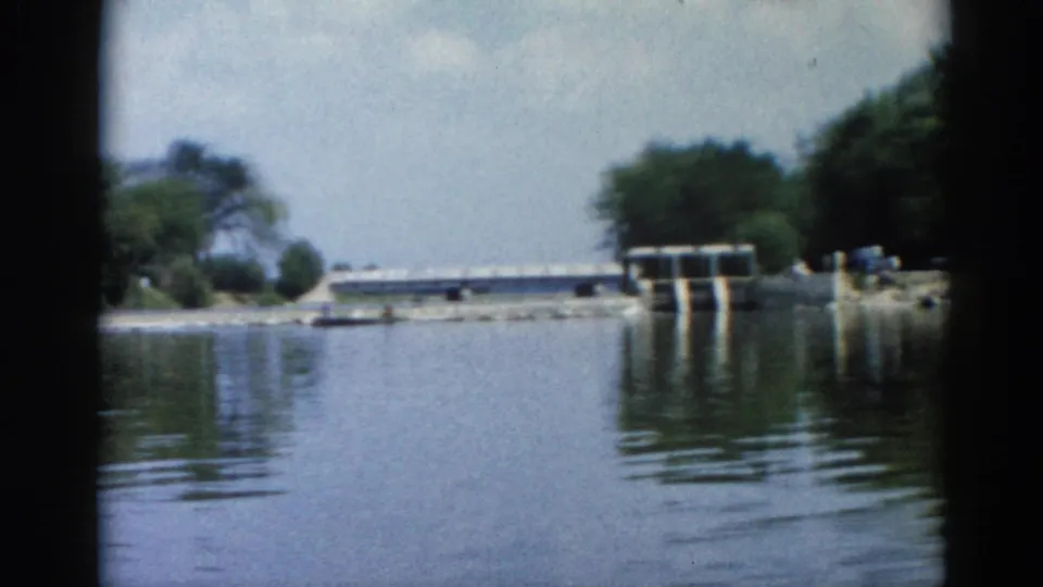 8mm home movie frame circa 1962 showing a calm Illinois river with a concrete dam or lock structure in the background and tree-lined banks under a hazy sky.