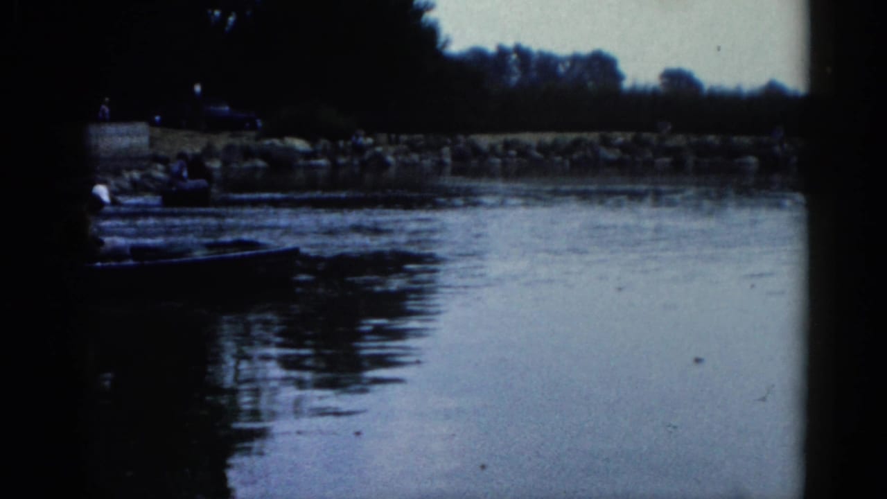 Rocky riverbank with large boulders; dark boats visible near the shore and a treeline in the distance under a pale sky.