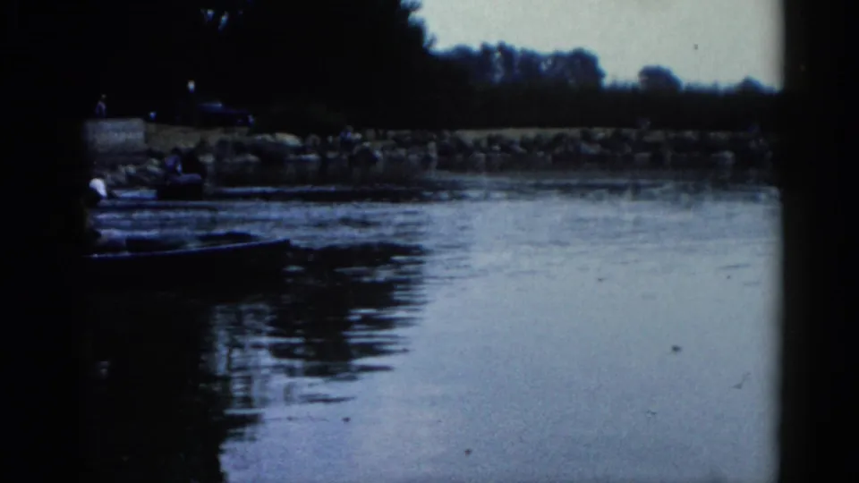 8mm home movie frame circa 1962 showing a rocky Illinois riverbank with dark boats near the shore and people faintly visible in the distance.