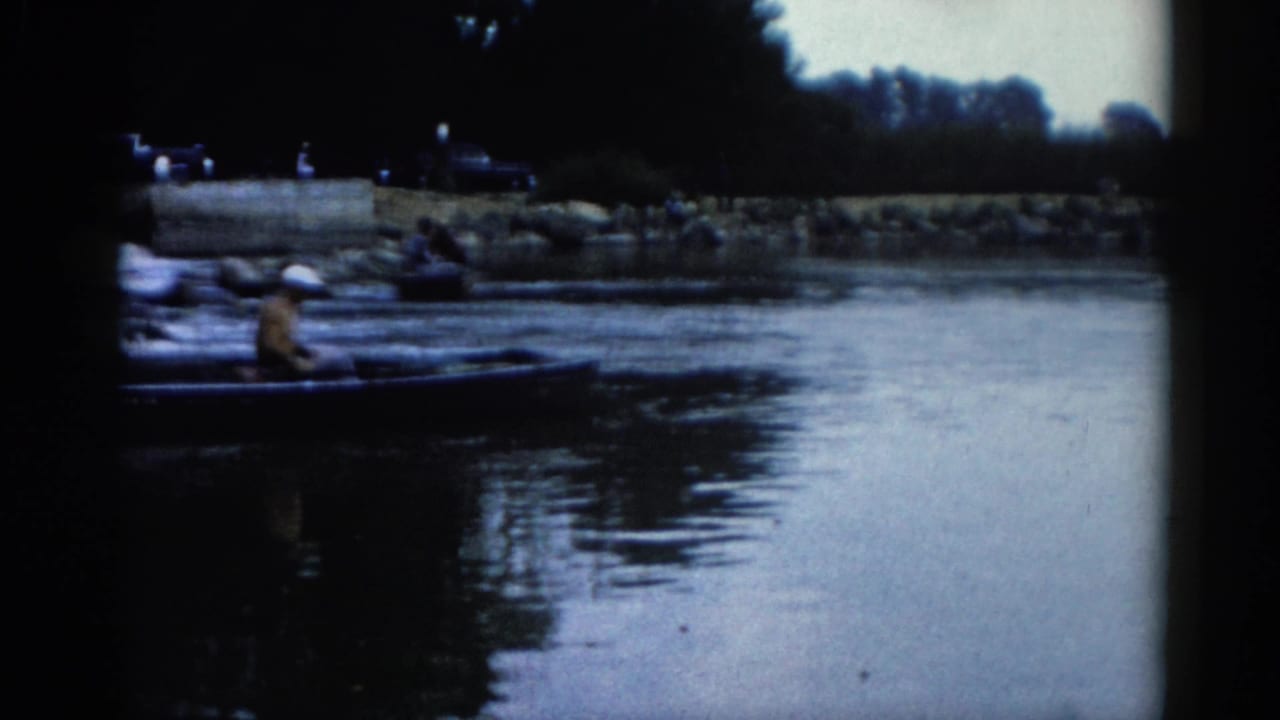 8mm home movie frame circa 1962 showing a person in a white hat seated in a small boat on an Illinois river, with a rocky bank and dark vehicle shapes visible behind.