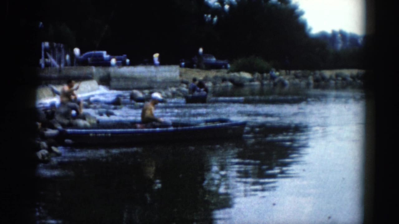 8mm home movie frame circa 1962 showing people gathered beside a small boat at a rocky Illinois riverbank, with what appear to be early-1960s cars parked on the shore.