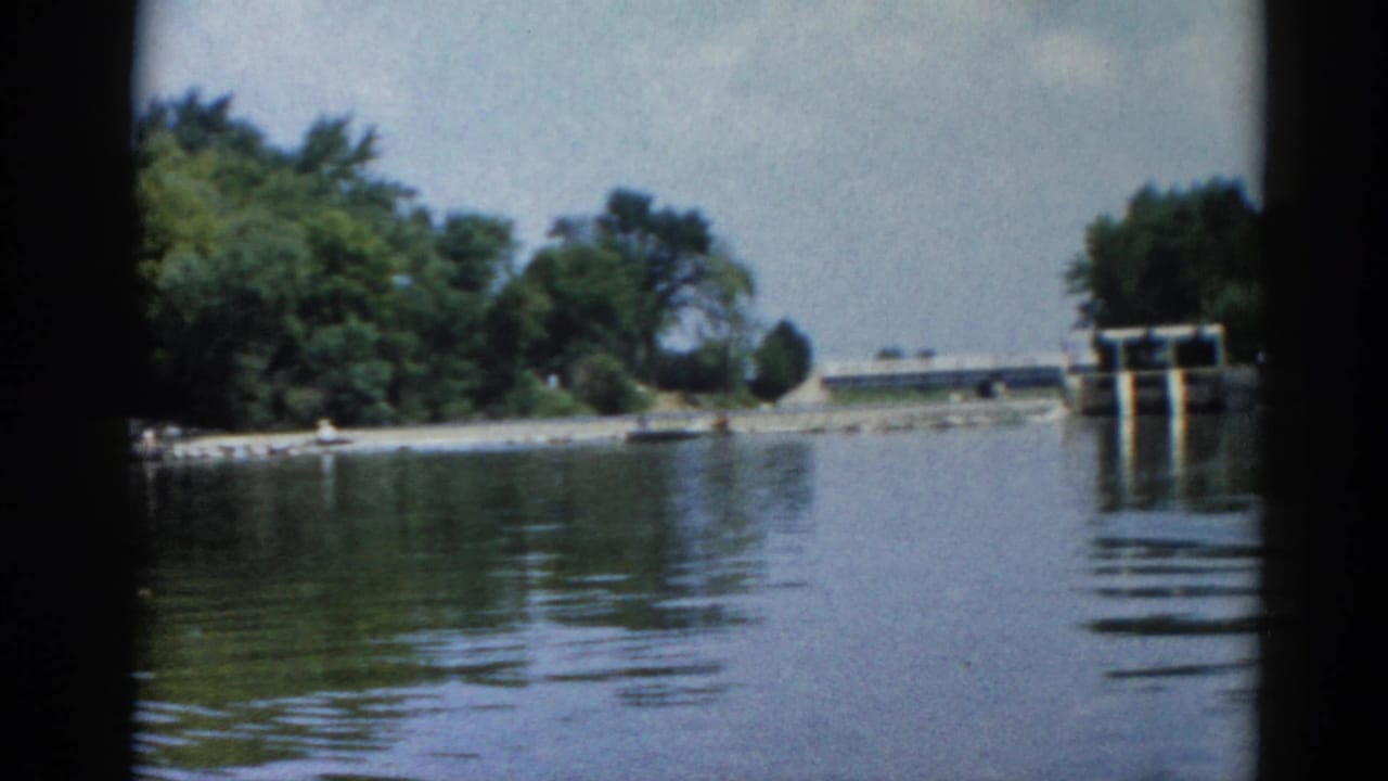 8mm film frame of a lake at water level, concrete dam structure visible in the background left, dense green treeline across the far bank.