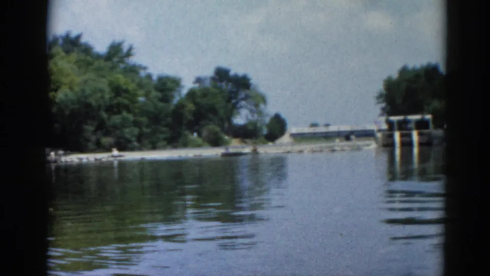8mm home movie frame, Illinois 1962: calm lake surface from a boat, concrete sluice-gate dam in background, full summer deciduous treeline and partly cloudy sky.
