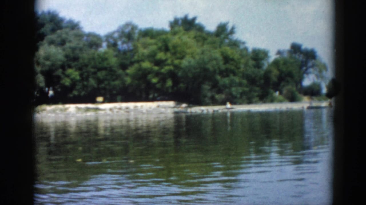 8mm home movie frame, Illinois 1962: rocky riprap shoreline along the right bank, calm lake water, and full summer deciduous canopy, shot from a moving boat.