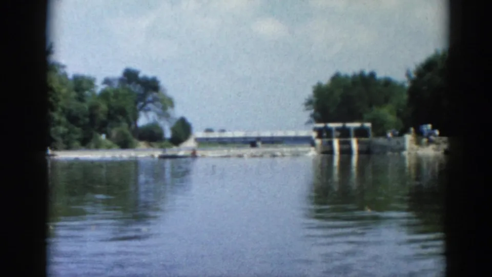 8mm home movie frame, Illinois 1962: concrete sluice-gate dam and rocky riprap bank seen from a moving boat on the lake, dense green treeline and partly cloudy sky beyond.