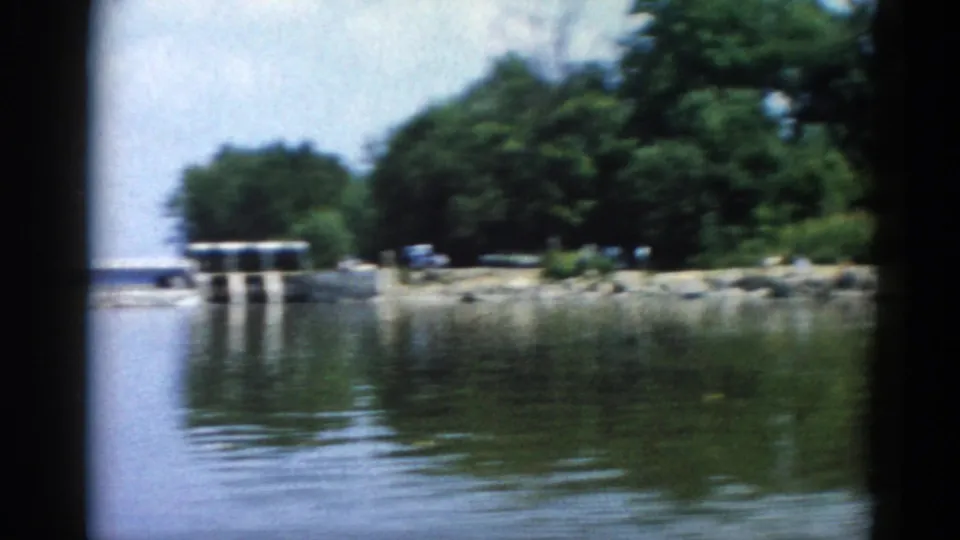 8mm home movie frame, Illinois 1962: concrete sluice dam with gate pillar supports and riprap bank, captured from a moving boat with summer treeline and blue-grey sky behind.