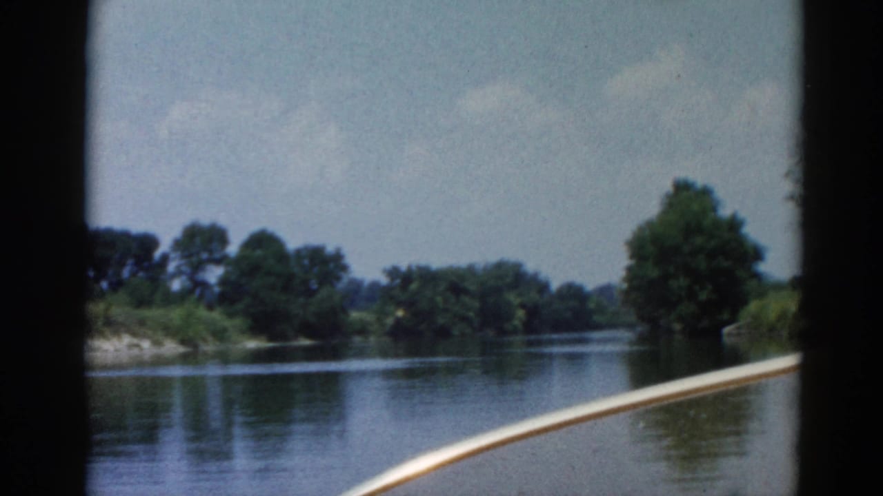 On-boat POV across a wide Illinois river, circa 1962; boat gunwale at lower right, dense summer treeline on far bank, 8mm color film.