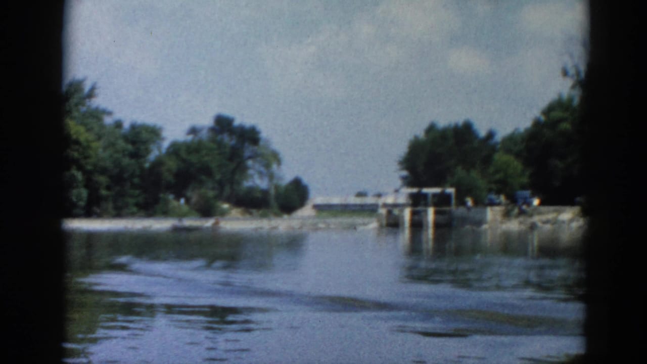 POV from a boat on an Illinois river, circa 1962, approaching a concrete navigation lock or dam; small figures and possible vehicles visible on shore.