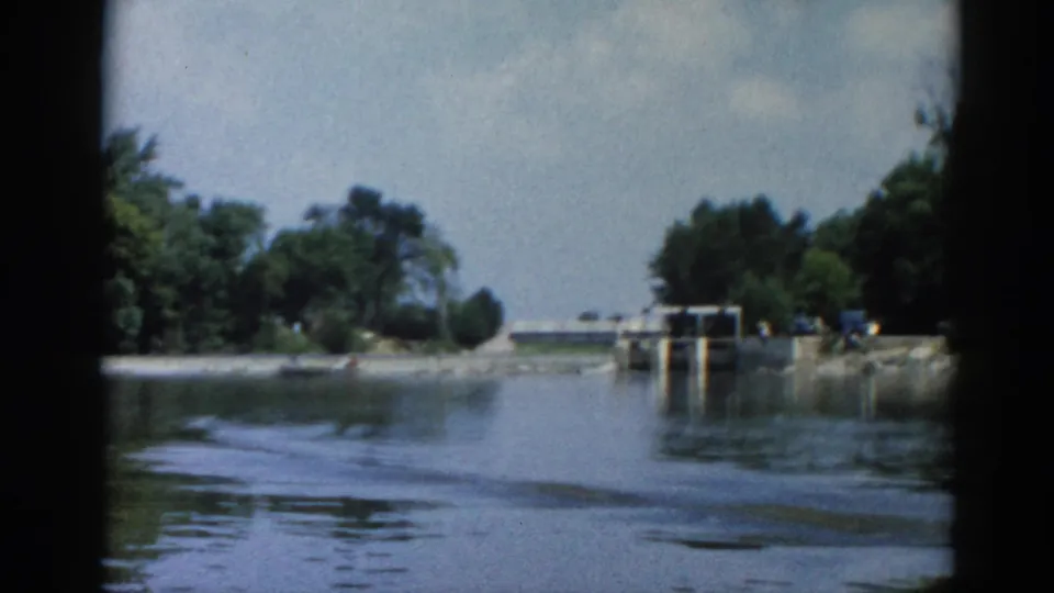 POV from a boat on an Illinois river, circa 1962, approaching a concrete navigation lock or dam; small figures and possible vehicles visible on shore.