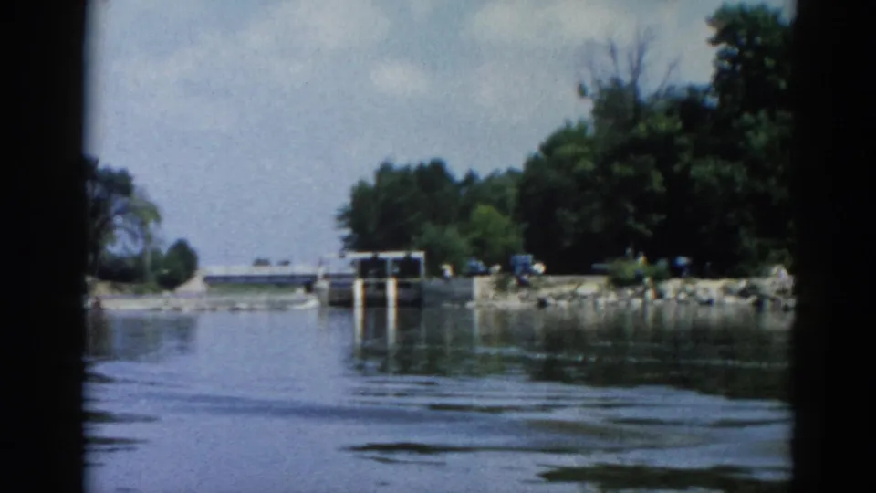Boat POV on an Illinois river, circa 1962, showing a concrete navigation lock with piers and rocky foreshore; small distant figures visible on shore.