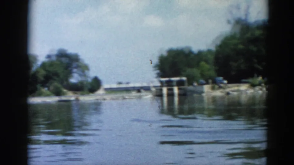 Boat POV on an Illinois river, circa 1962; concrete lock or dam structure with rocky foreshore, small distant figures visible, dense treeline above.