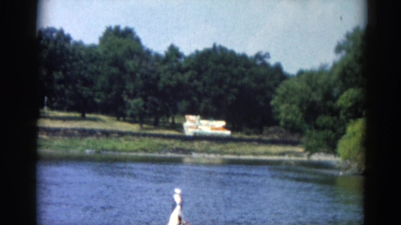 Person in white hat standing at motorboat bow on a wide river; colorful boat visible on far wooded shore.