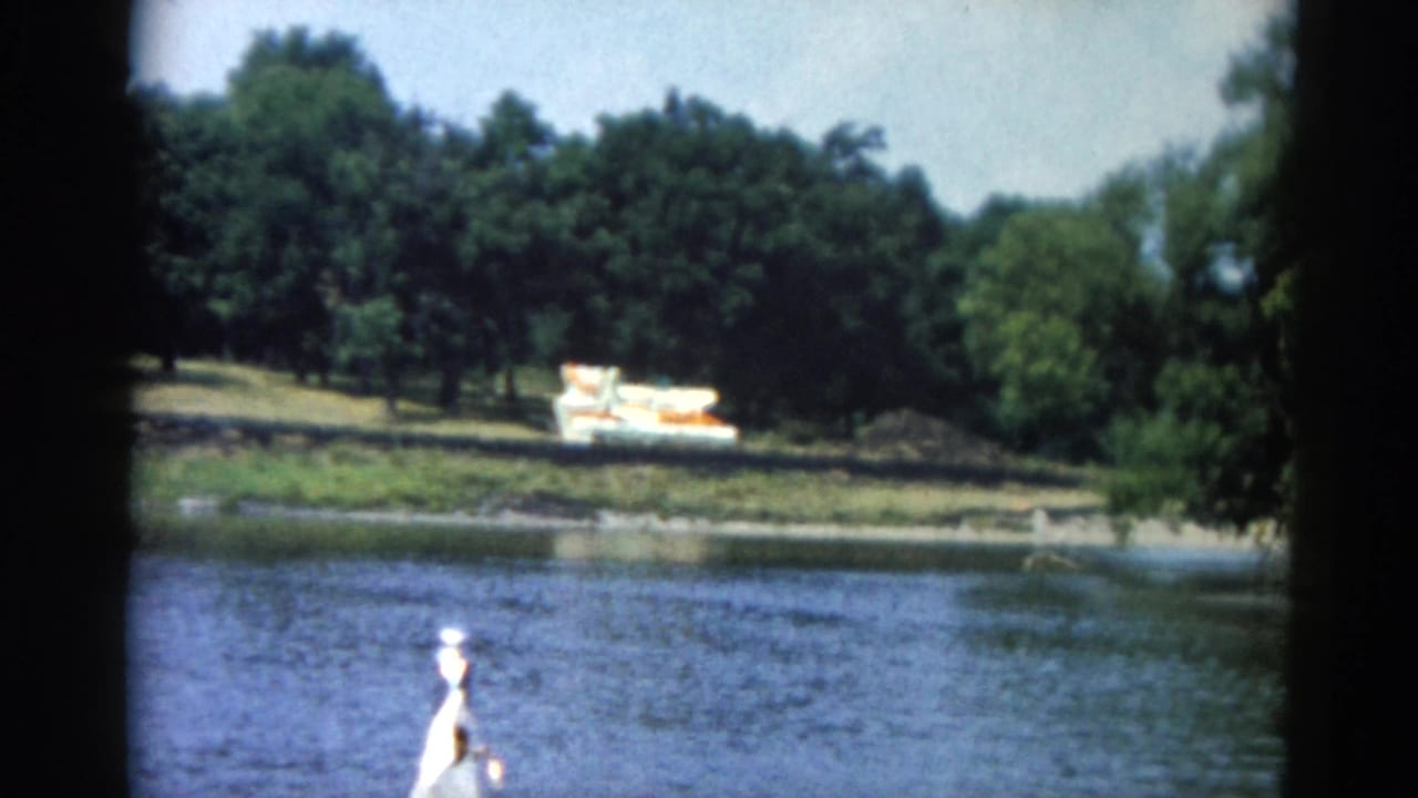 Continuing river journey from motorboat; figure in white hat at bow, colorful boat still visible on wooded far shoreline.