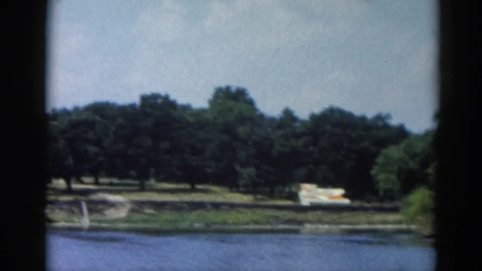 8mm footage of a calm Illinois lake circa 1962; open water in the foreground and a tree-lined bank with boats on the opposite shore under a partly cloudy sky.