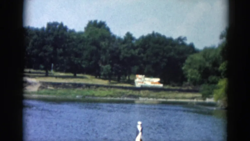 8mm Illinois lake footage circa 1962; open water, wooded opposite bank with boats on the grass, and a small white element at the waterline whose nature requires human review.