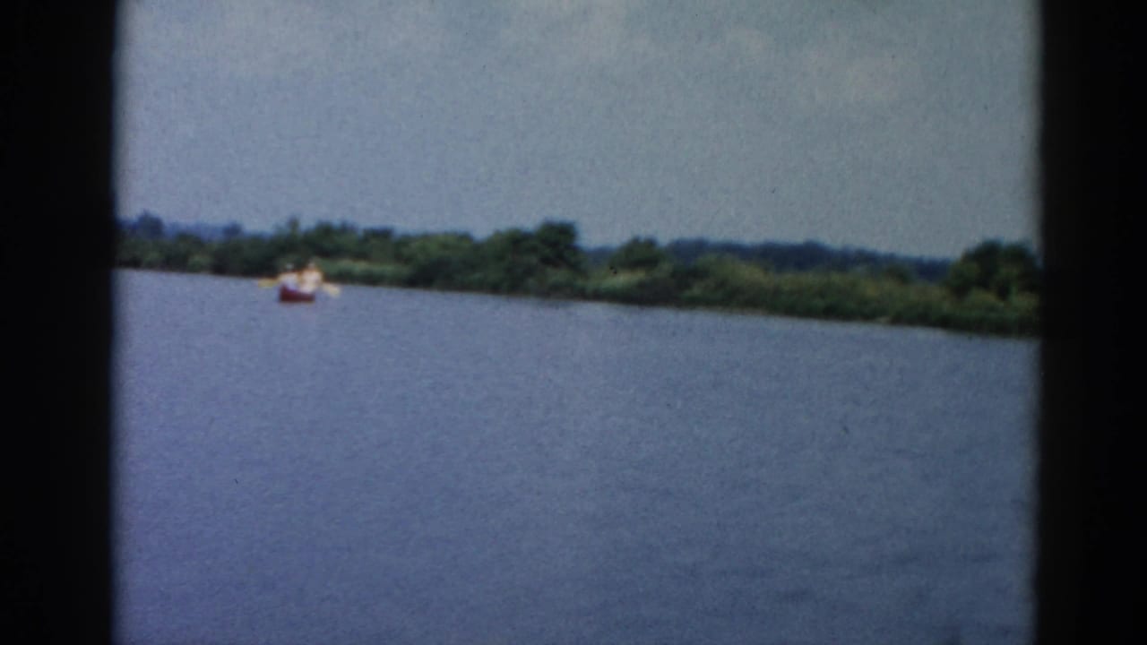 8mm home movie frame from 1962 Illinois showing a broad river, a small red boat with occupants near a lush deciduous treeline, under an overcast sky.