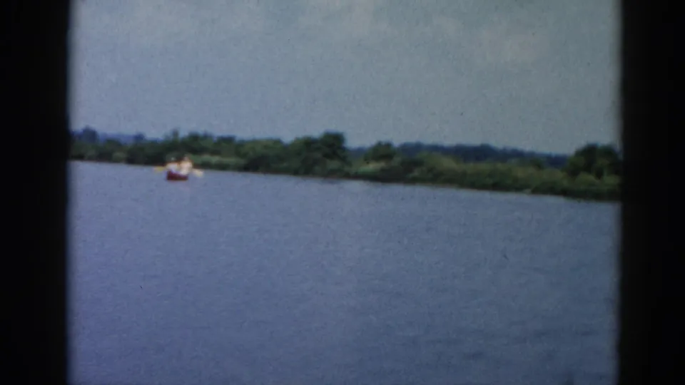 8mm home movie frame from 1962 Illinois showing a broad river, a small red boat with occupants near a lush deciduous treeline, under an overcast sky.