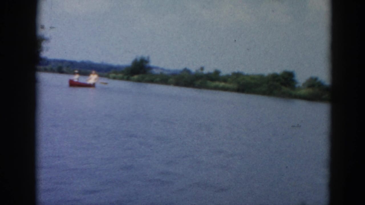 8mm frame circa 1962 Illinois showing a red boat with an occupant in white crossing a broad river, wooded far shore behind under pale sky.
