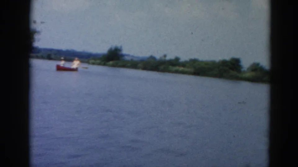 8mm frame circa 1962 Illinois showing a red boat with an occupant in white crossing a broad river, wooded far shore behind under pale sky.