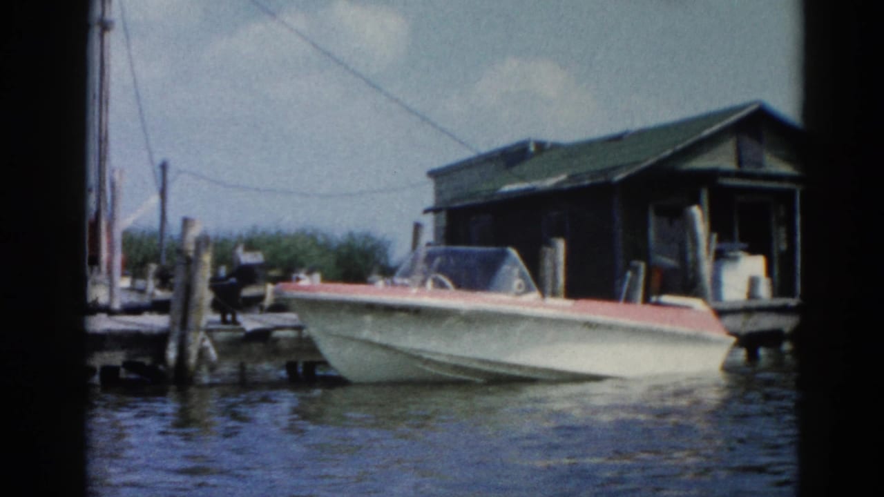 White speedboat with red trim moored at wooden dock pilings; dark boathouse behind; 8mm home movie.