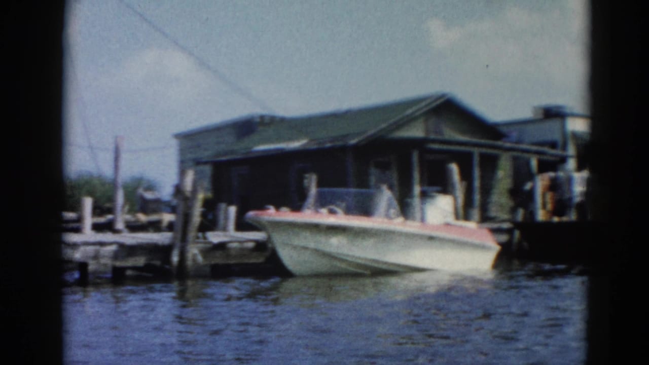 White speedboat with red trim at wooden dock; dark boathouse and overhead power lines visible; 8mm home movie.