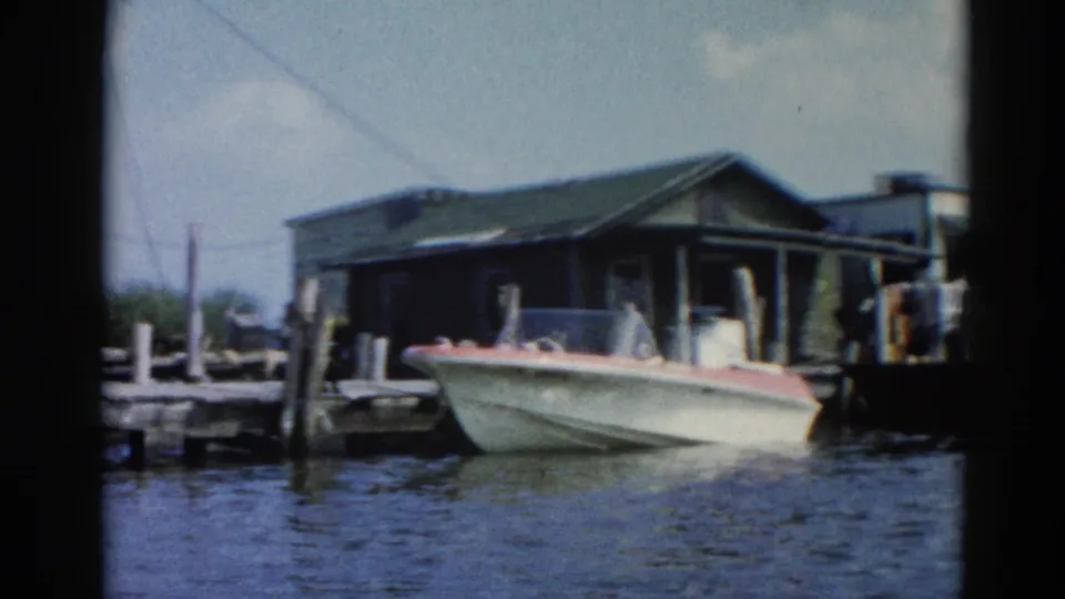 White speedboat with red trim moored at a wooden dock, dark boathouse with green roof and utility lines overhead, Illinois waterfront circa 1962.