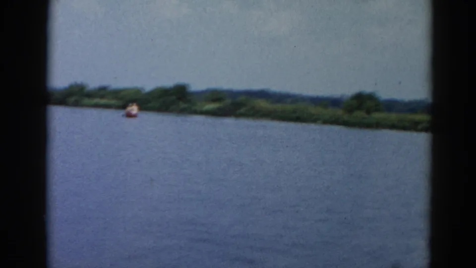 Wide shot of open lake water with treeline on the far shore and a small figure in a distant boat, Illinois, circa 1962, 8mm home movie.
