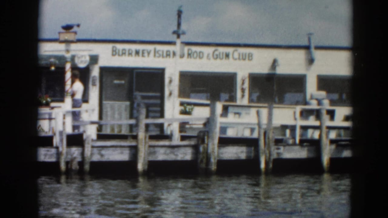 Wooden pier with white 'Blarney Island Rod & Gun Club' building; a figure stands near the entrance; calm water in foreground.