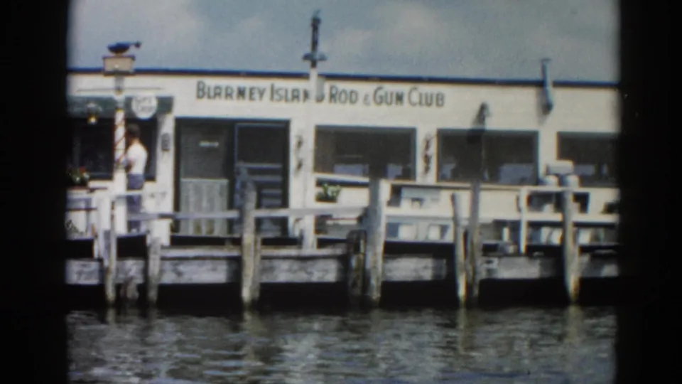 8mm home movie frame of the Blarney Island Rod & Gun Club on a wooden pier, Illinois circa 1962, with a figure near the entrance over calm rippling water.