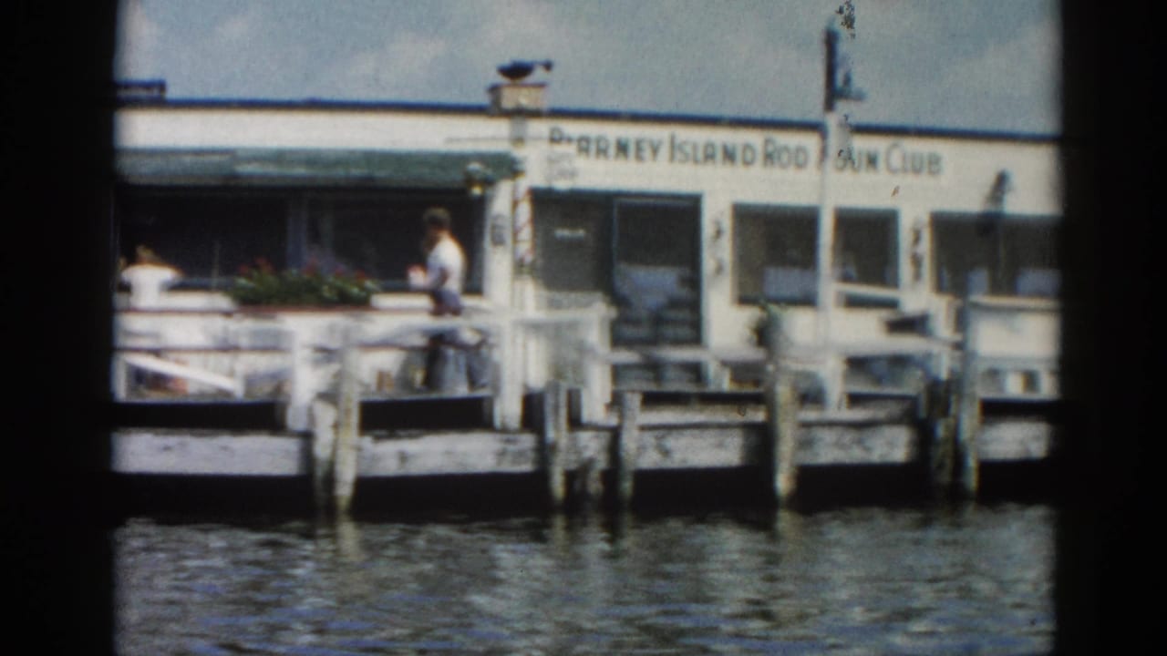 Same Blarney Island Rod & Gun Club pier from water level; a figure visible on the right deck section; rippling water in foreground.