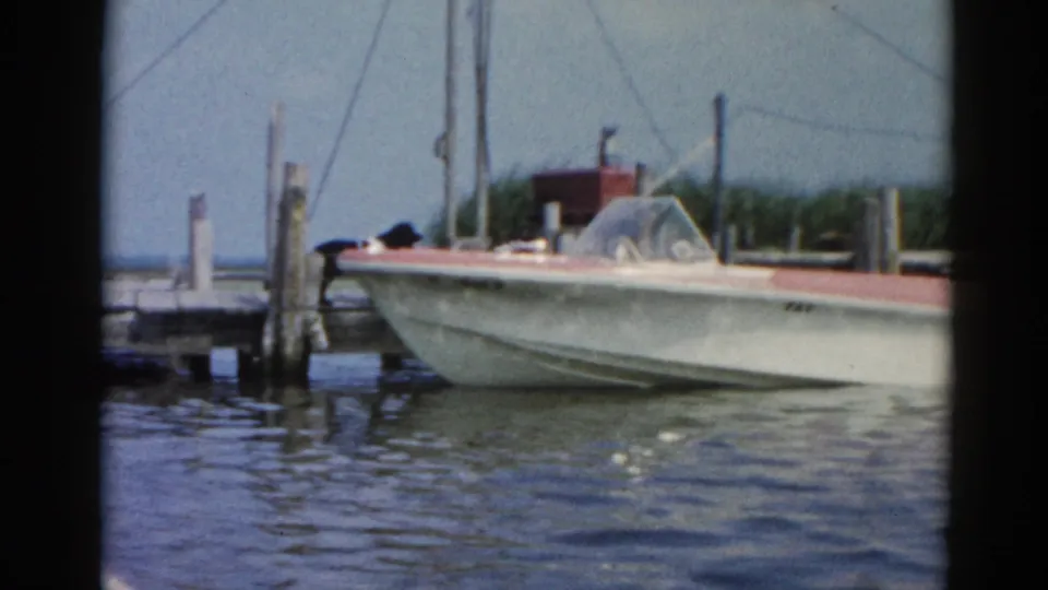 White speedboat with red trim moored at a Midwestern dock, Illinois waterway circa 1962; marsh reeds and hazy sky visible in background.