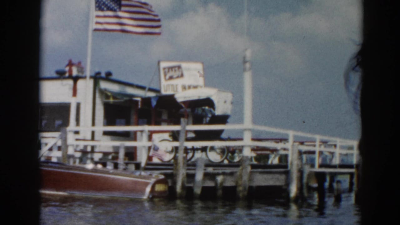 Vintage wooden motorboat moored at a dock with white railings; American flag on a pole above; overcast sky.