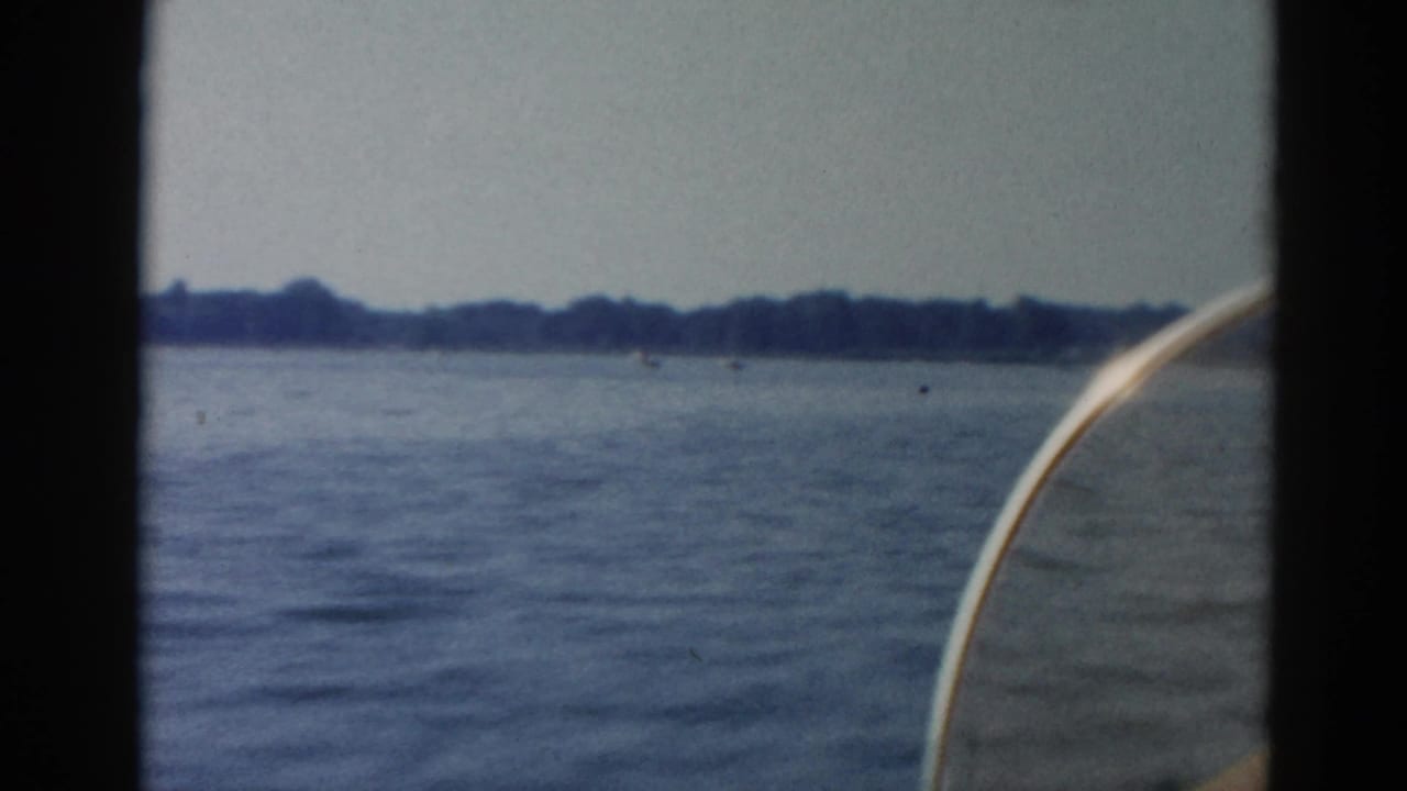 Lake view through a window frame, open water with distant tree-lined shore and overcast sky, bow of a boat visible at right edge.