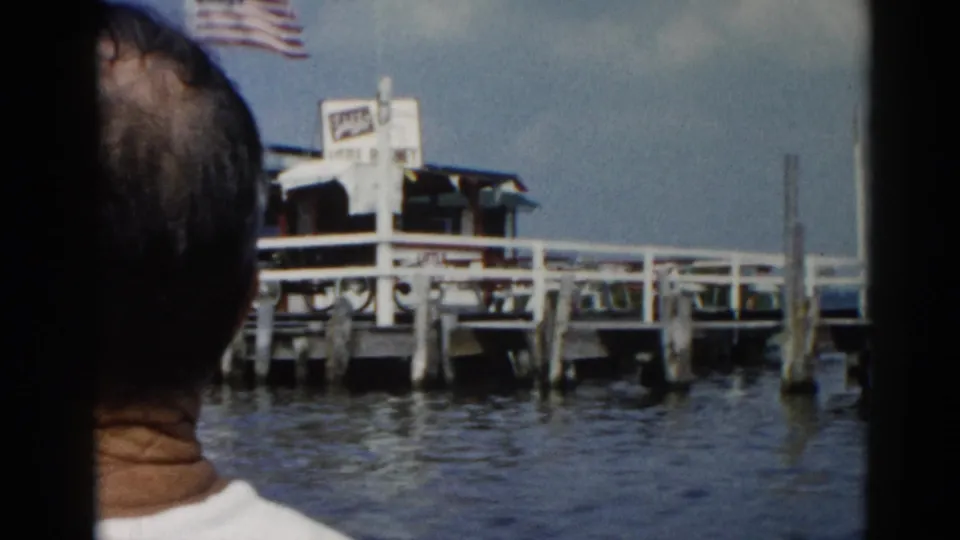 8mm home movie circa 1962 from a boat approaching an Illinois lake marina; person's back visible in foreground with American flag and dock signage ahead.