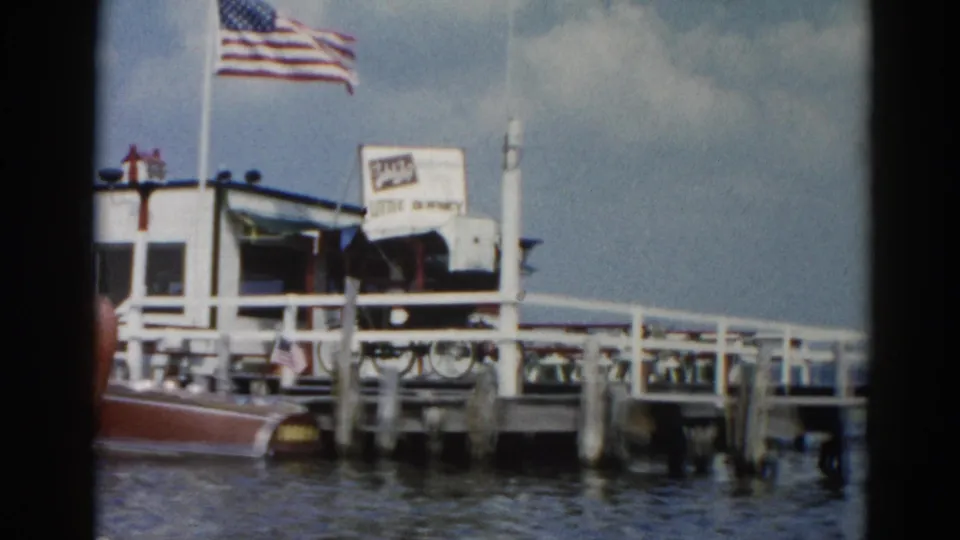 8mm home movie circa 1962 showing an Illinois lake marina dock with a flying American flag, 'Little Ducky' sign, wooden pilings, and a classic wooden motorboat.