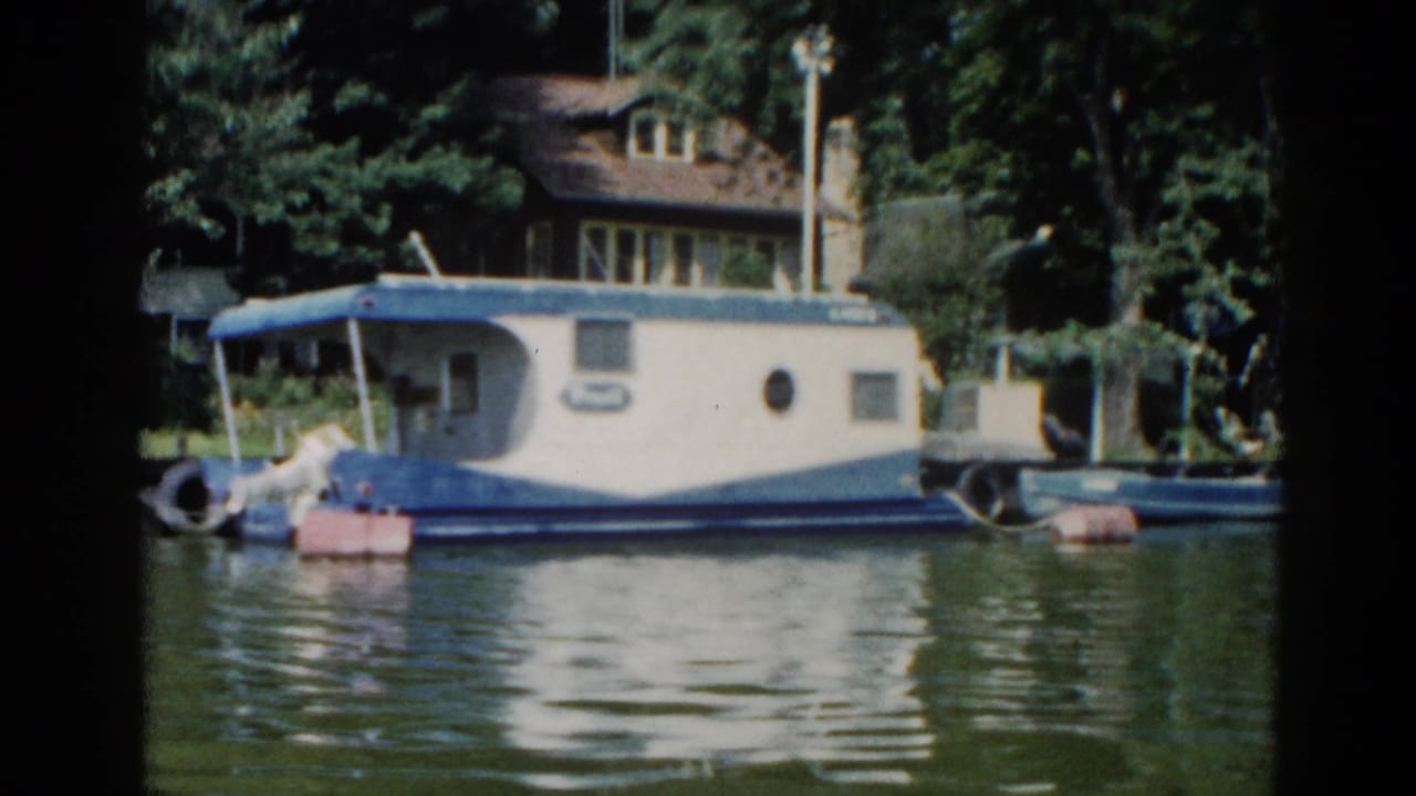White and blue houseboat moored at a dock, brick cottage and trees in background.