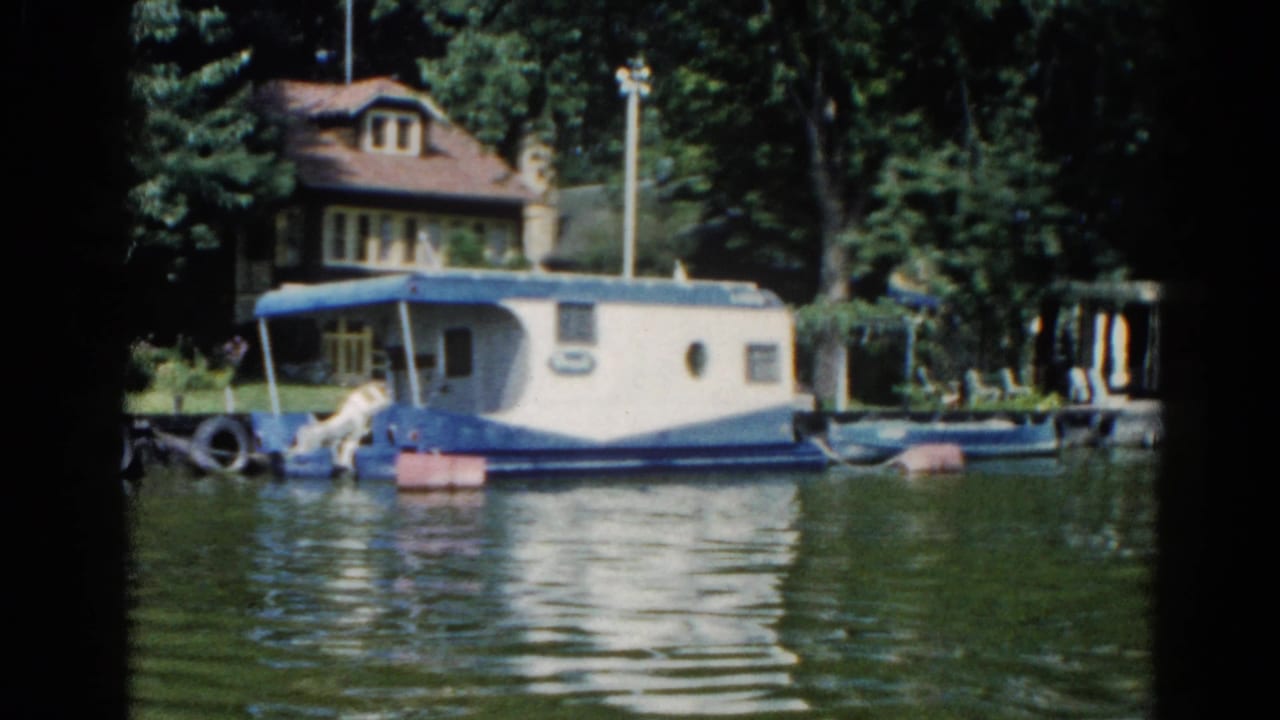 Second view of Illinois houseboat and dock, circa 1962, showing blue canopy and porthole windows with lakeside cottage and treeline behind.