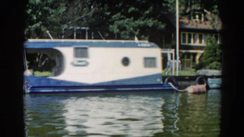 8mm footage circa 1962 Illinois showing a blue-and-white houseboat with porthole windows docked along a tree-lined shore, residential building behind