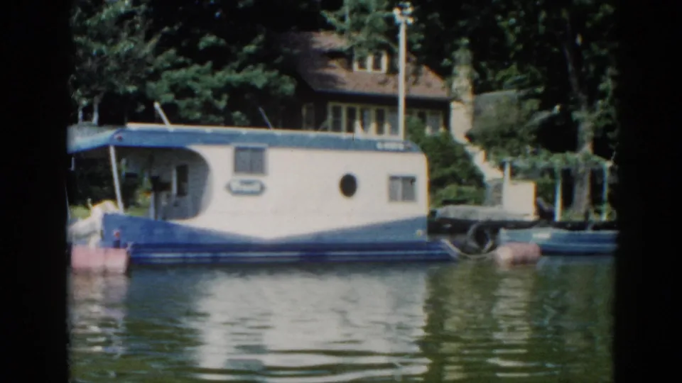 8mm footage circa 1962 Illinois with a blue-and-white houseboat in the foreground, tree canopy, and a residential building with large windows behind