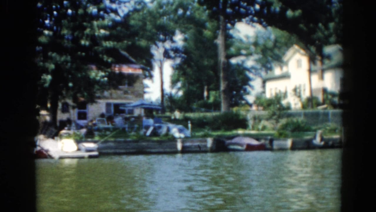 1962 Illinois home movie shot from a motorboat showing a residential shoreline with a white house, docked boats, and summer foliage