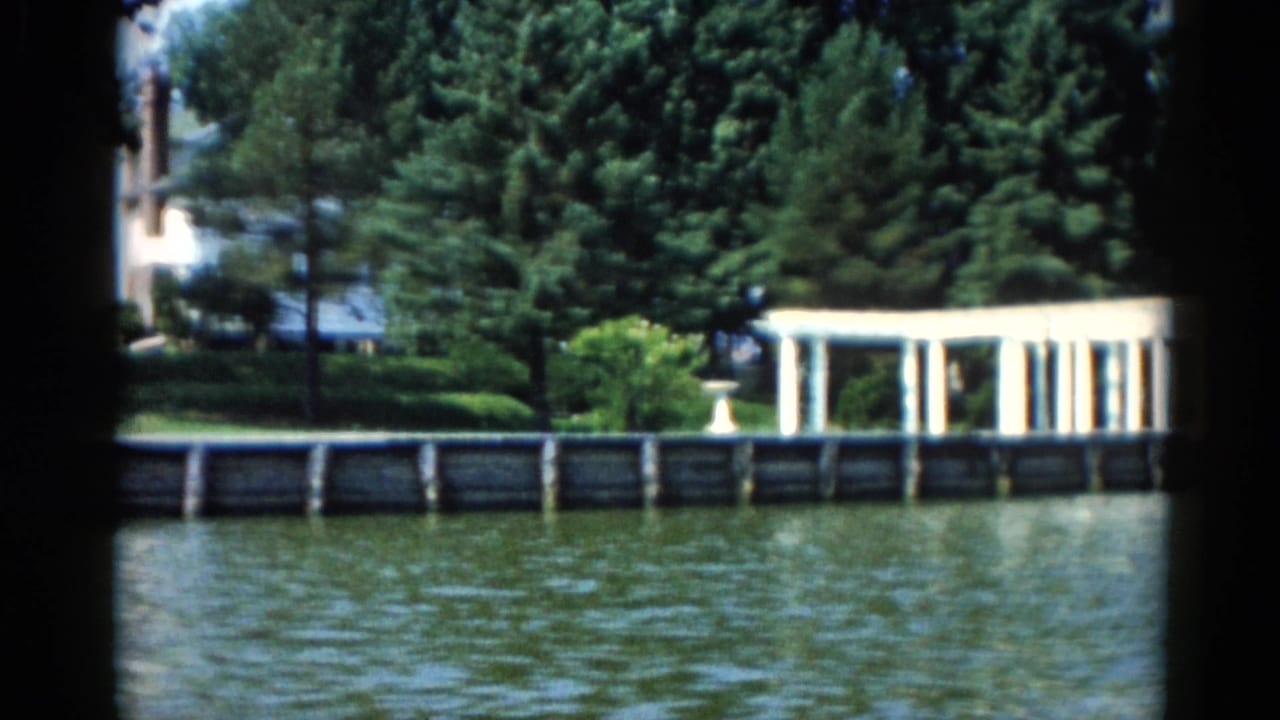 Riverside view of a white columned pergola amid dense trees and manicured lawn, seen from the water on 8mm film.