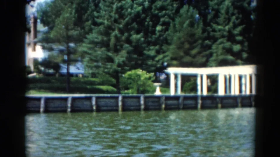 1962 Illinois riverfront showing a white columned pergola, manicured hedges, and seawall along a calm river, on 8mm home movie film.